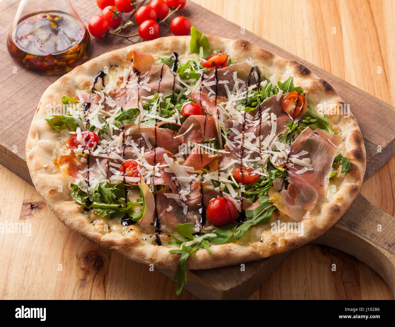 Pizza with dried ham, rocket leaves, tomato and Parmesan cheese on a cutting board. Stock Photo