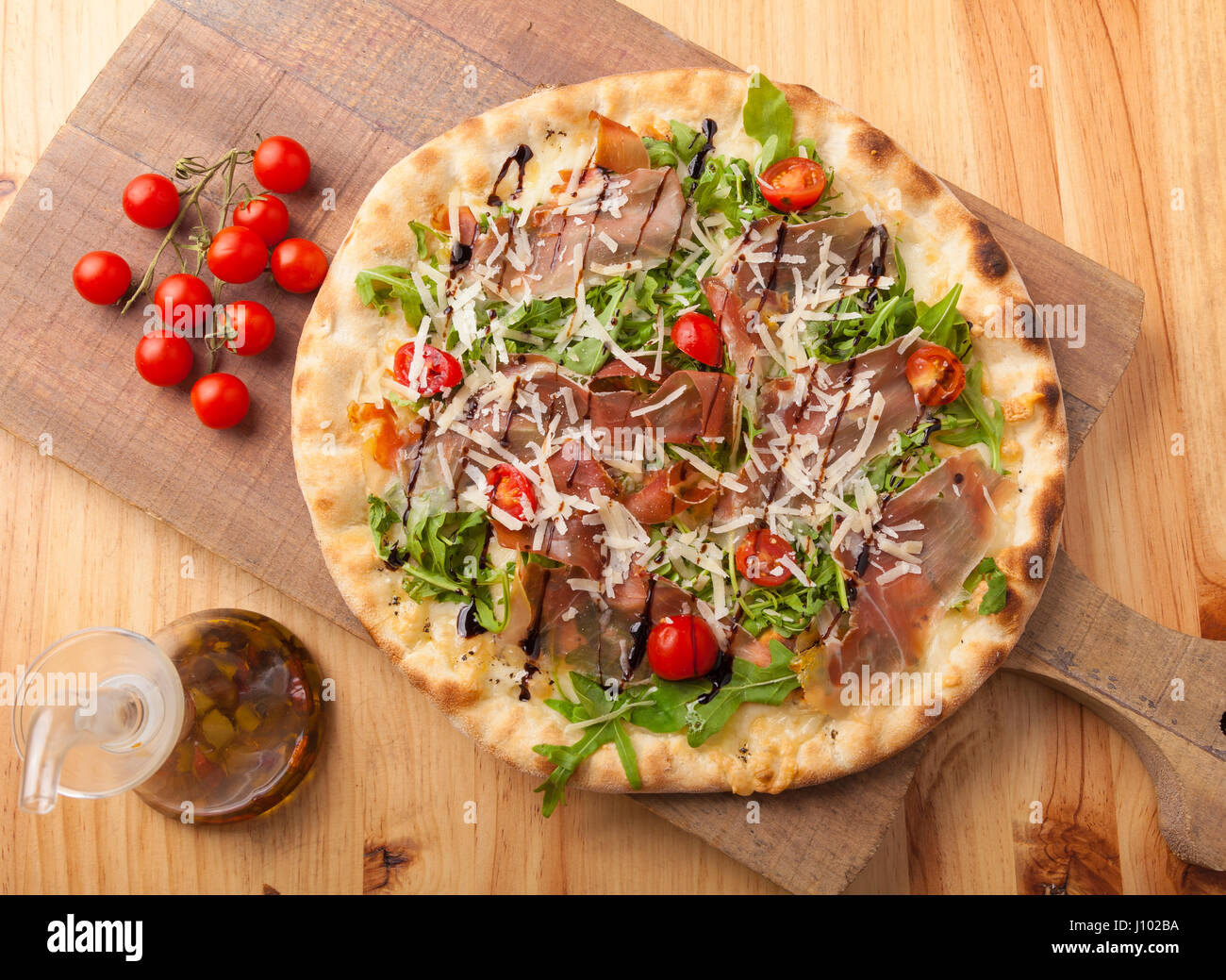Pizza with dried ham, rocket leaves, tomato and Parmesan cheese on a cutting board. Stock Photo