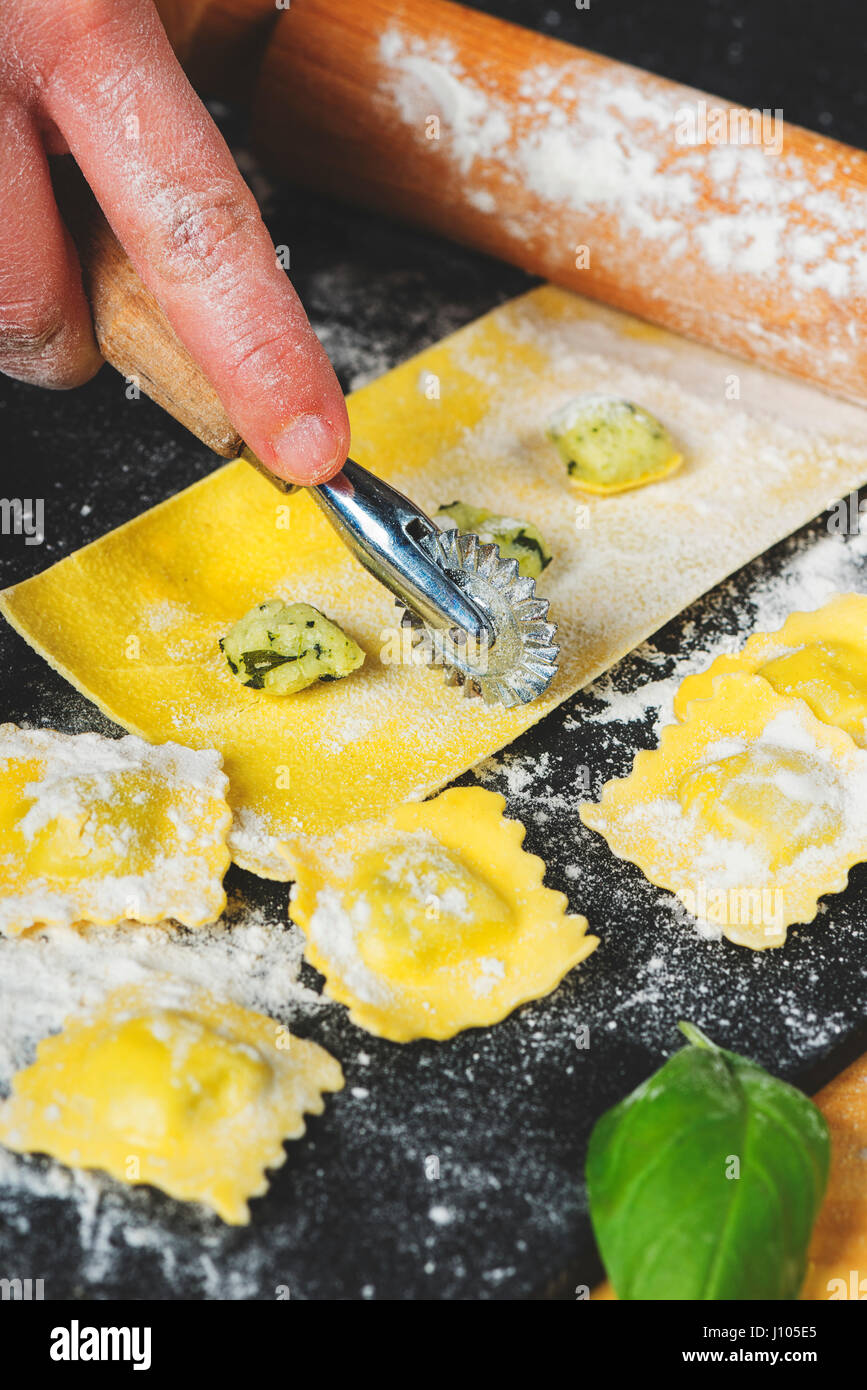 Preparing ravioli in the kitchen with tools and ingredients : dough, flour, eggs, stuffing, cutter, roller, board. Cutting squares to finish prepare. Stock Photo