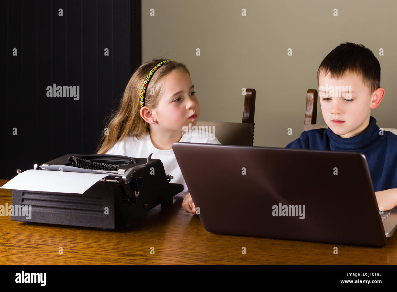 Girl angry because she has to work on an old typewriter while her brother is working on a modern laptop Stock Photo