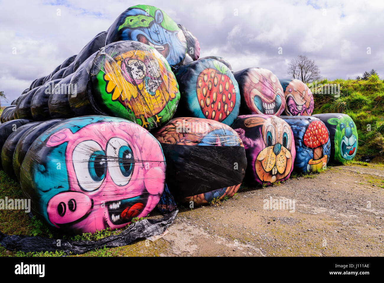 Haybales stacked at the side of a road with various colourful faces and objects painted on them. Stock Photo