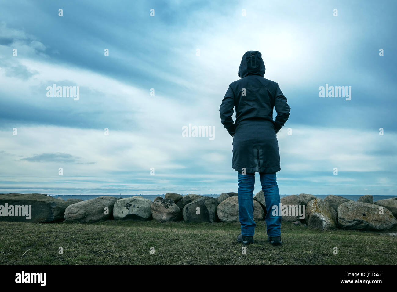 Lonely hooded female person from behind standing at seashore and looking into distance on a cold winter day, concept of waiting, anticipation, hope an Stock Photo