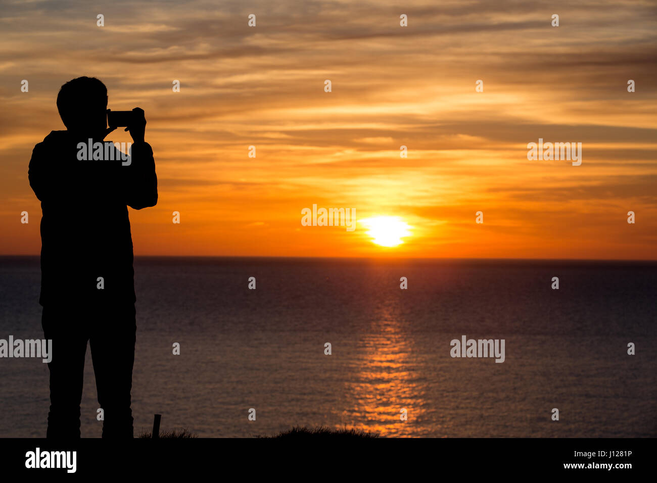 Silhouette of man taking a picture of a coastal sunset with his mobile phone Stock Photo
