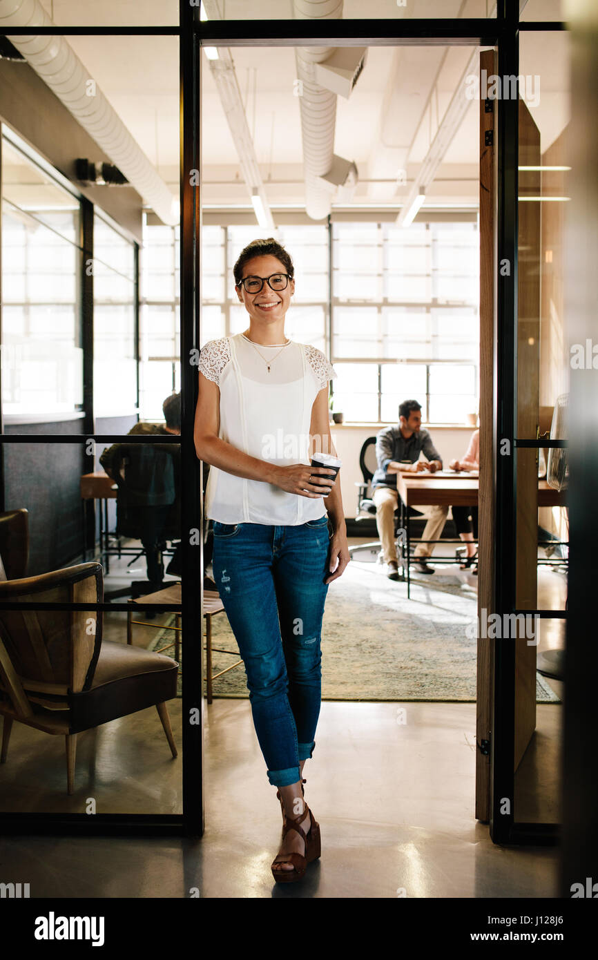 Full length portrait of young woman at office doorway holding a coffee. Relaxed female executive having coffee break with colleagues in background. Stock Photo