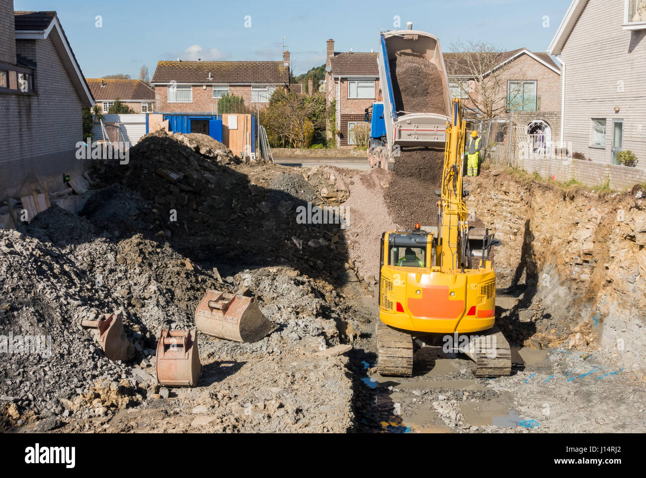 Excavator is digging a house foundation in a residential area while a dumper truck is unloading construction gravel, sand and crushed stones on the co Stock Photo