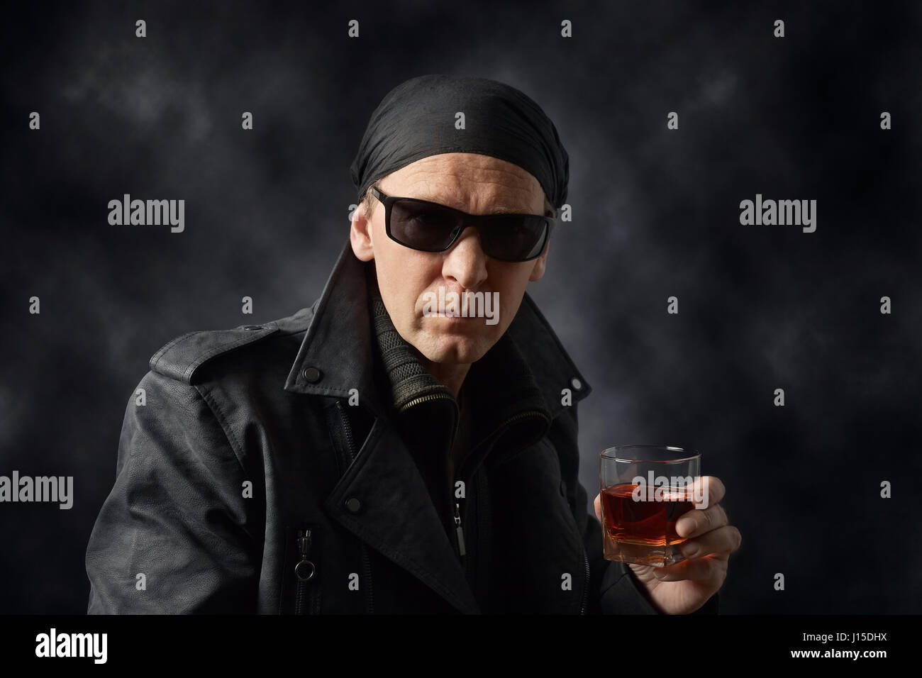 Portrait of a middle-aged man in a black leather jacket , sunglasses and black bandana .Rocker with glass of whiskey on black background. Stock Photo