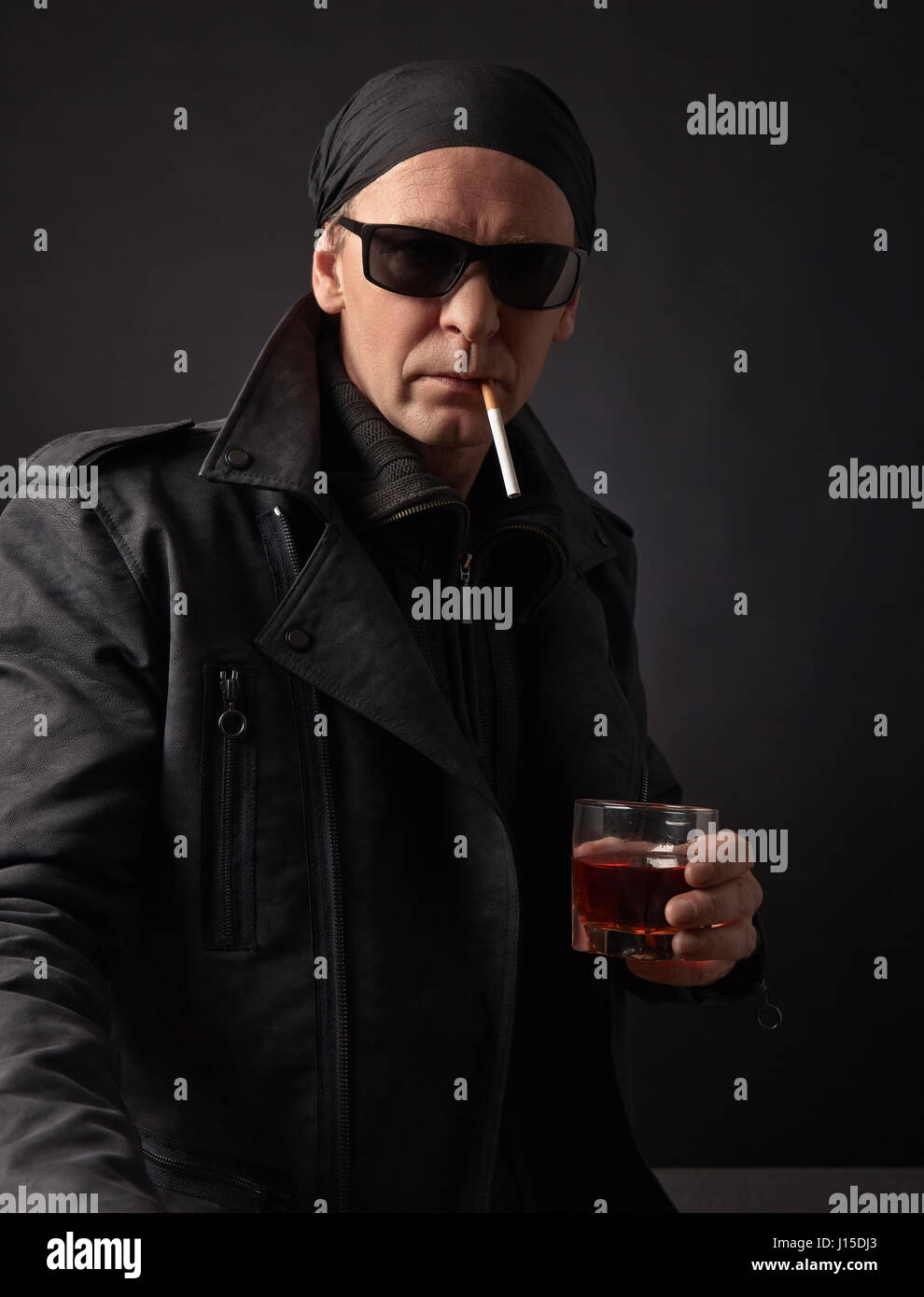 Portrait of a middle-aged man in a black leather jacket , sunglasses and black bandana .Rocker with glass of whiskey and cigarette on black background Stock Photo