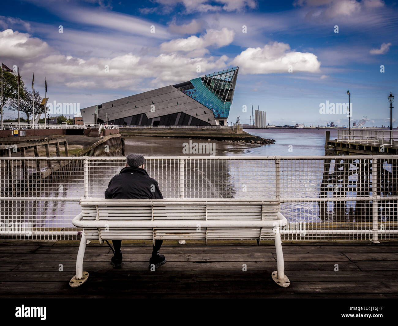 Old man wearing flat cap sitting on bench looking over water to The Deep, Visitor attraction, Hull, UK. Stock Photo