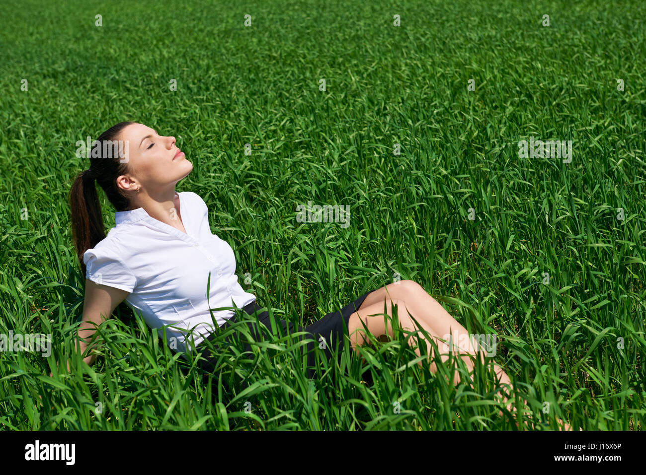 Business woman relaxing in green grass field outdoor under sun. Beautiful young girl dressed in suit resting, spring landscape, bright sunny day Stock Photo