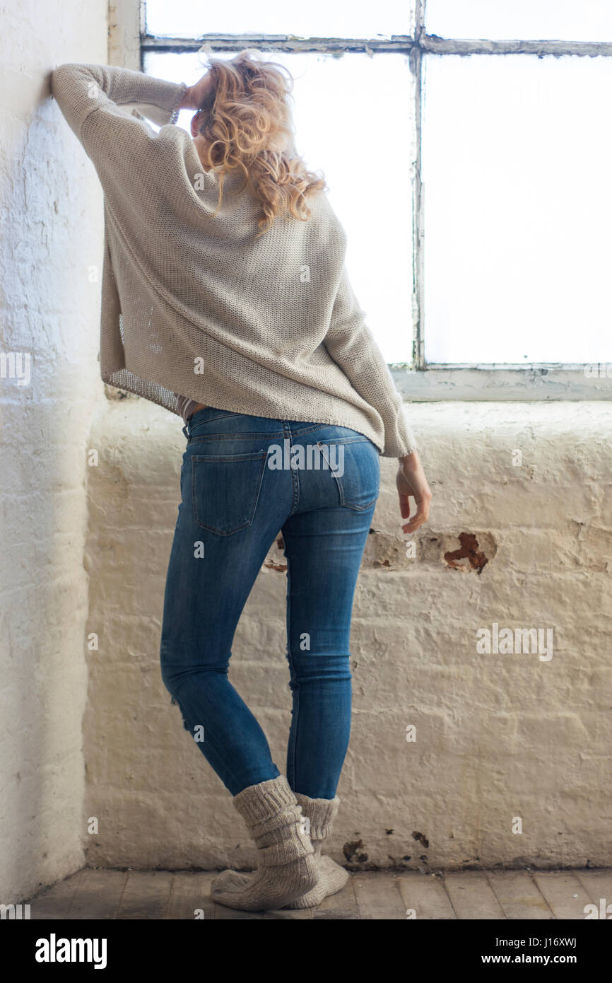 Slim woman wearing jeans standing by the window leaning against the wall Stock Photo