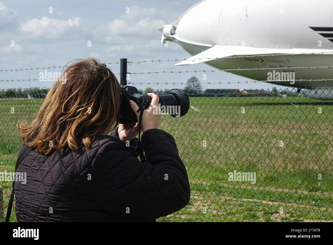 Cardington, Bedfordshire, UK. 18th Apr, 2017. The Hybrid Air Vehicles Airlander 10 is moored to the new Mobile Mooring Mast (MMM), a female photographer shoots through the mesh fence at the airfield. An Auxiliary Landing System (ALS) has been added which allows the aircraft to land safely at a greater range of landing angles. Photo Credit: Mick Flynn/Alamy Live News Stock Photo
