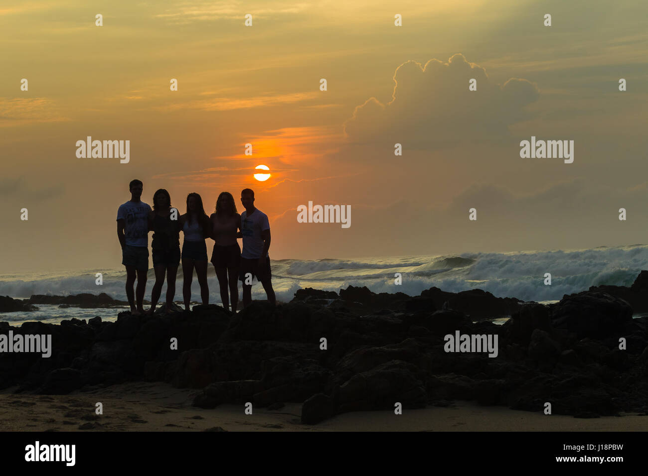 Friends girls boys teenagers morning beach ocean sunrise silhouetted along rocky ocean coastline. Stock Photo