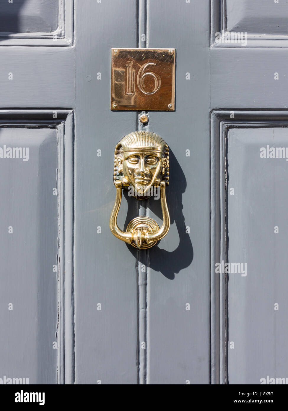 Gleaming brass door knocker in the style of an Egyptian sphinx with a brass number plate above of sixteen on a mid grey door Stock Photo