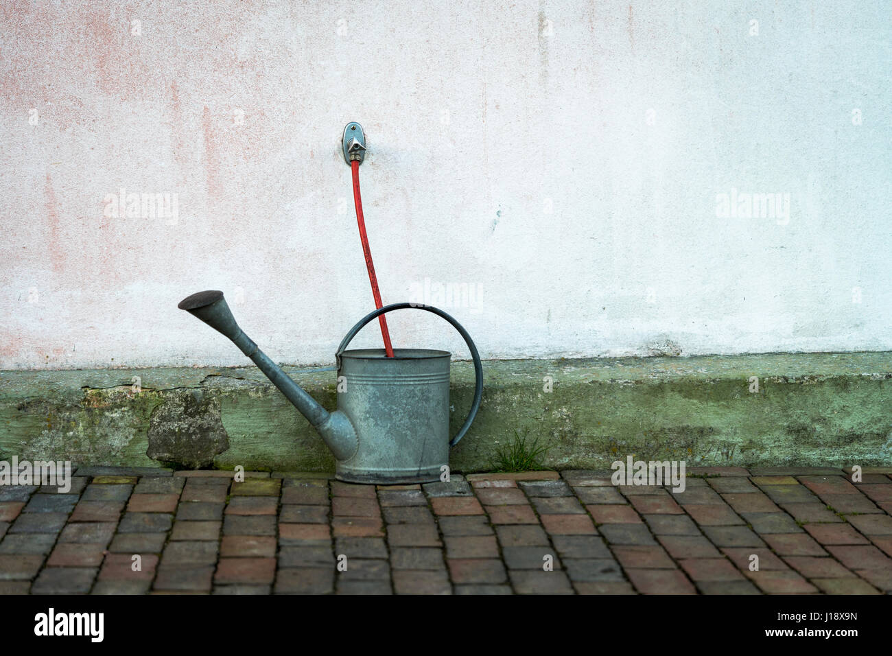 Urban gardening with grey watering can and red garden hose against weathered wall Stock Photo