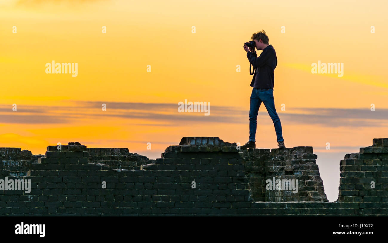 Photographer standing on a wall in the evening holding a camera taking a photo, with an orange sky from the low evening sun. Stock Photo