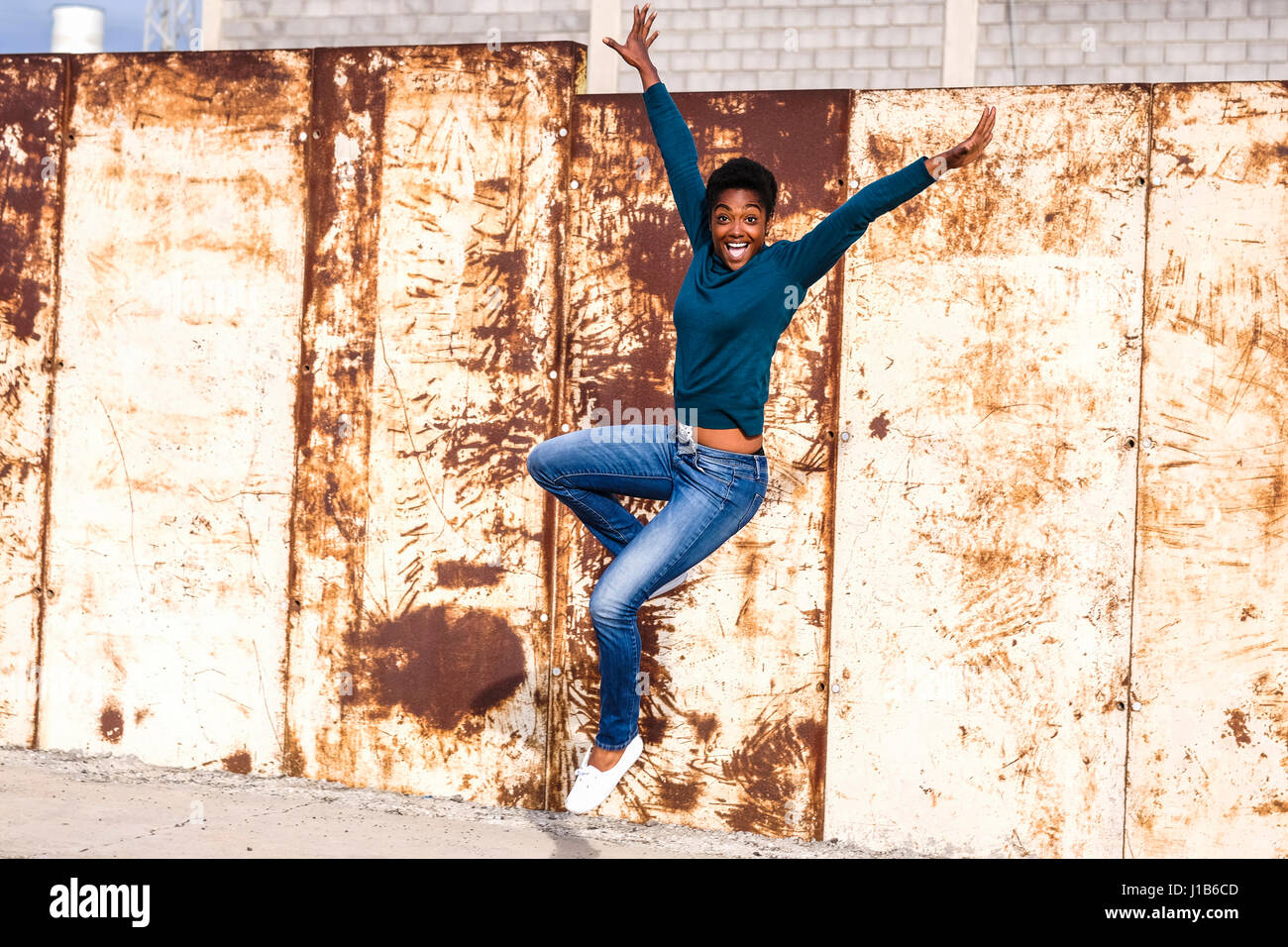 African American woman jumping for joy near rusty metal wall Stock Photo