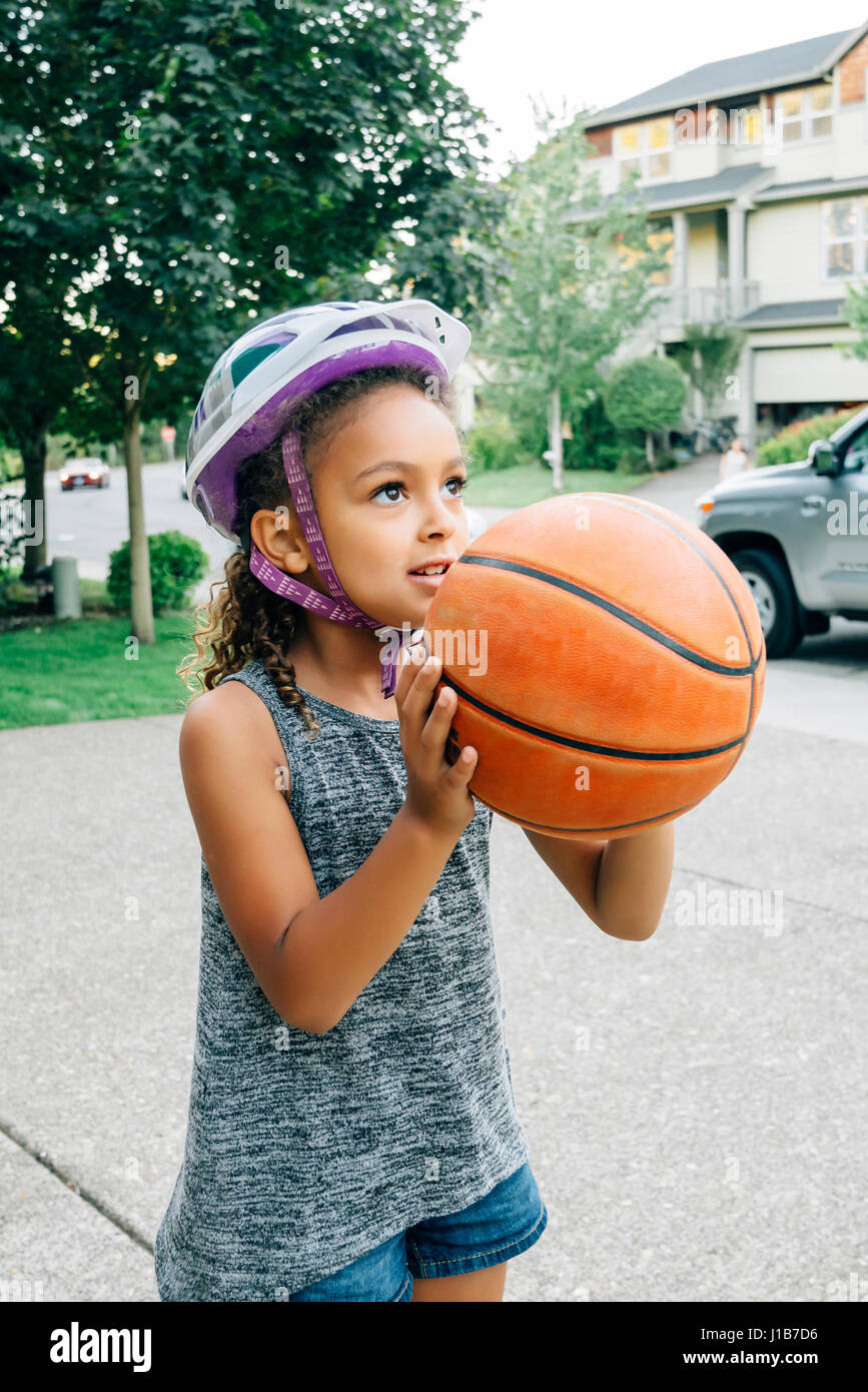 Mixed Race girl wearing helmet playing basketball in driveway Stock Photo
