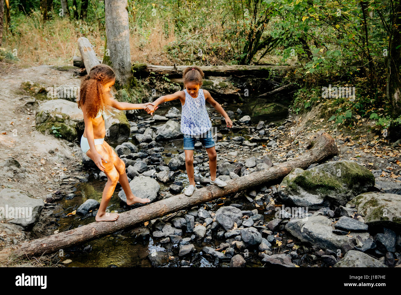 Mixed Race sisters holding hands on log over forest stream Stock Photo