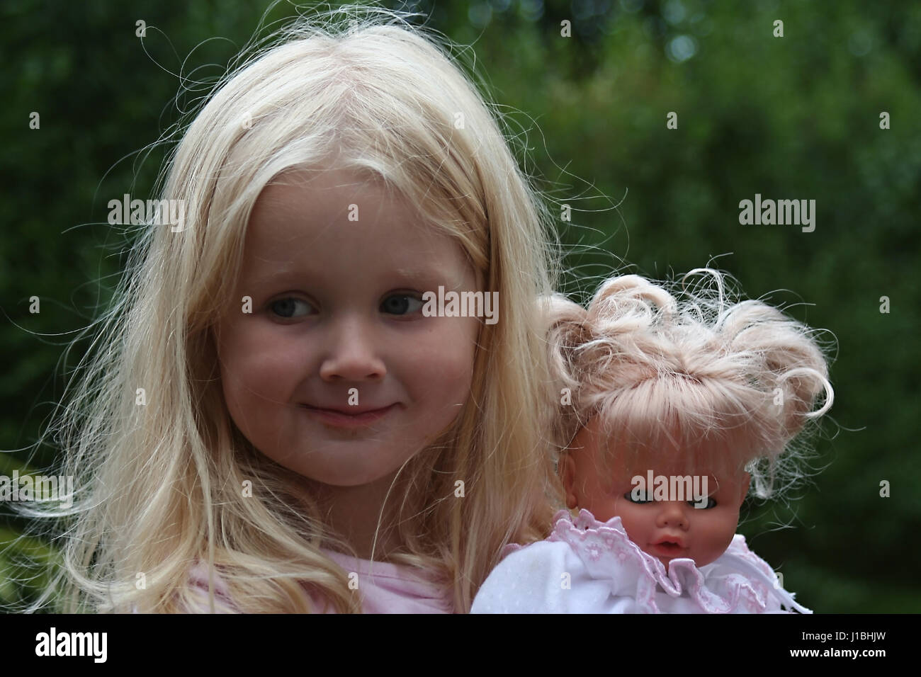 Little blonde girl, child playing with her doll in the garden blurred background, love concept, children toys, joy fun playtime, little girl doll Stock Photo