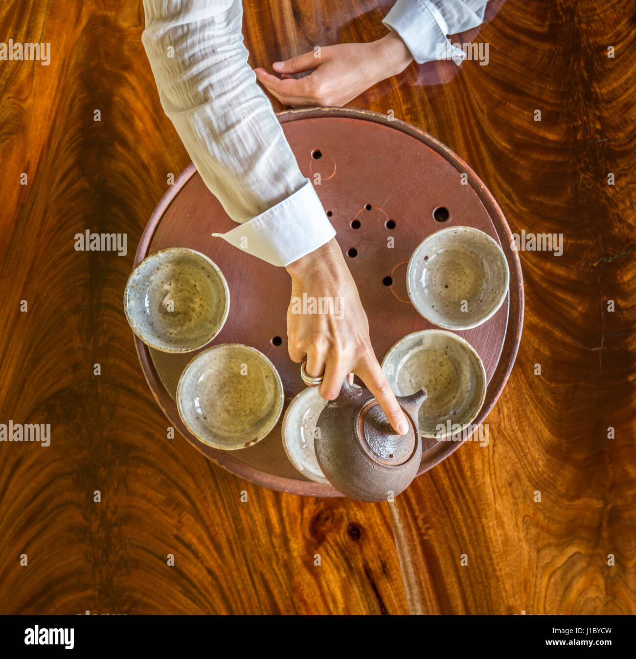 Top down view from above of Sarah Heddin's hands pouring tea into a ceramic pot on a tray with tea cups. Stock Photo