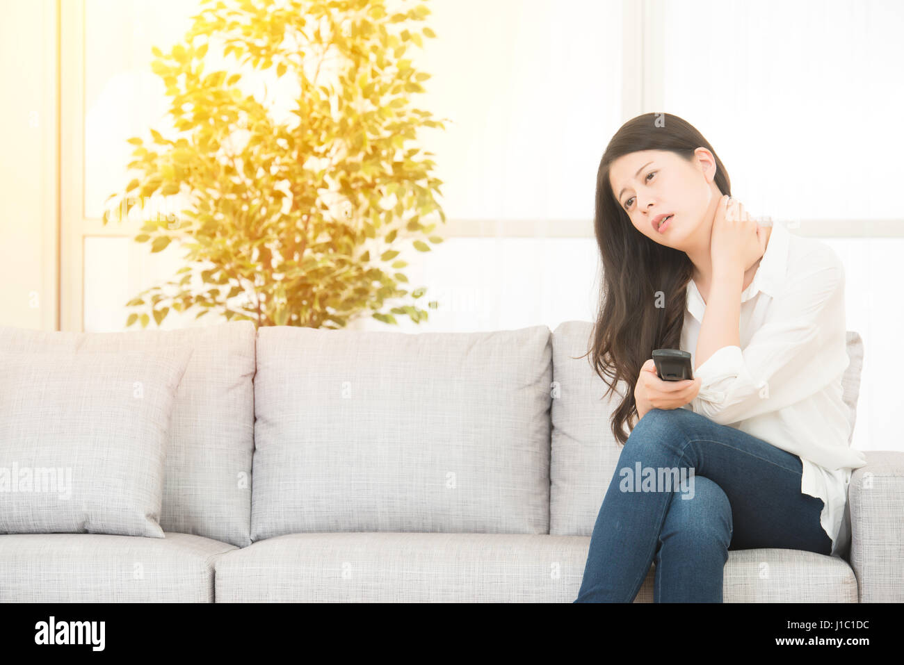 beautiful woman suffering from painful neck sitting on cosy sofa watching TV. lifestyle and health concept. mixed race asian chinese model. Stock Photo