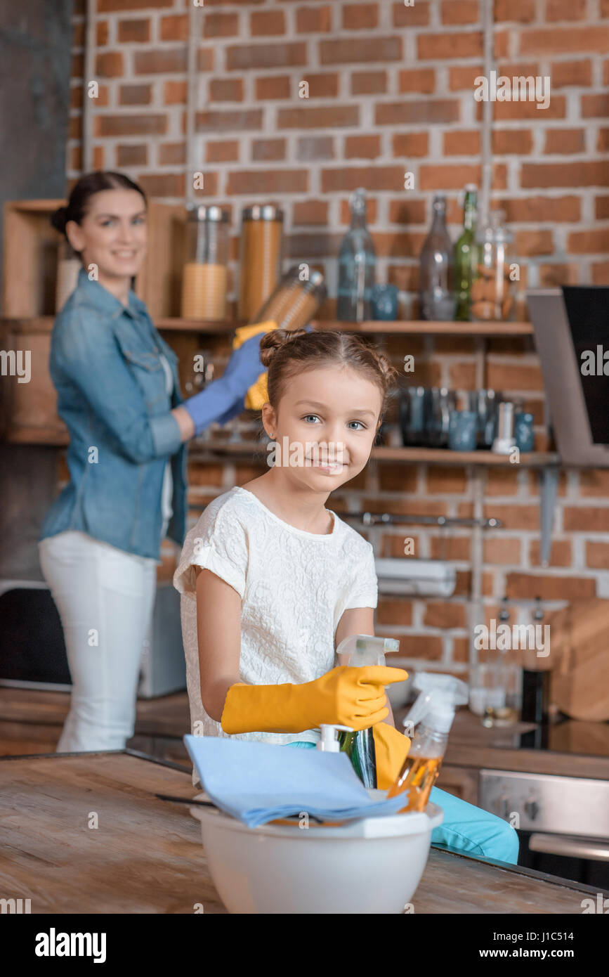 smiling girl in rubber gloves with cleaning supplies and mother behind ...