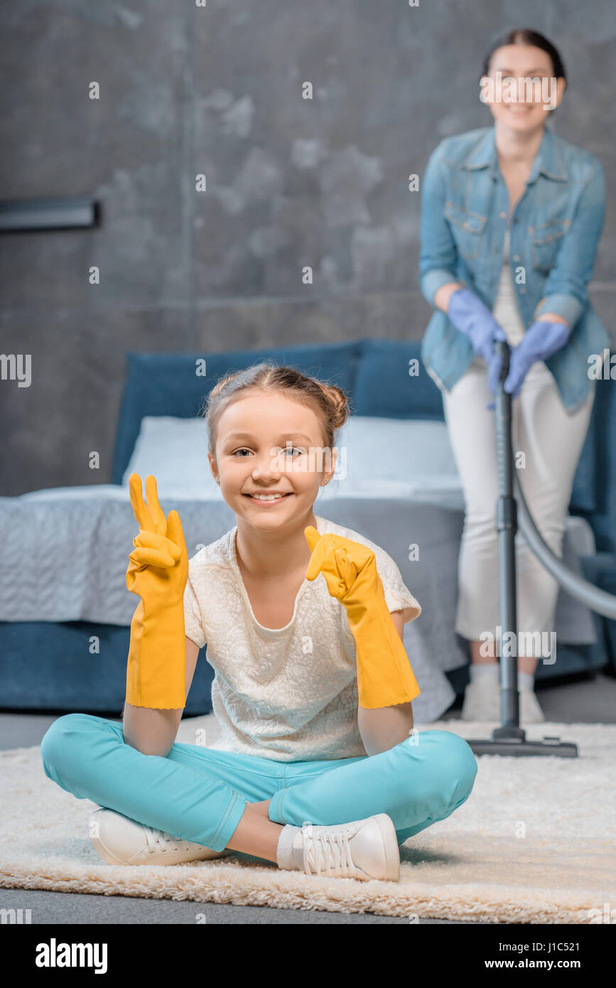 Mother with vacuum cleaner looking at happy daughter in rubber gloves