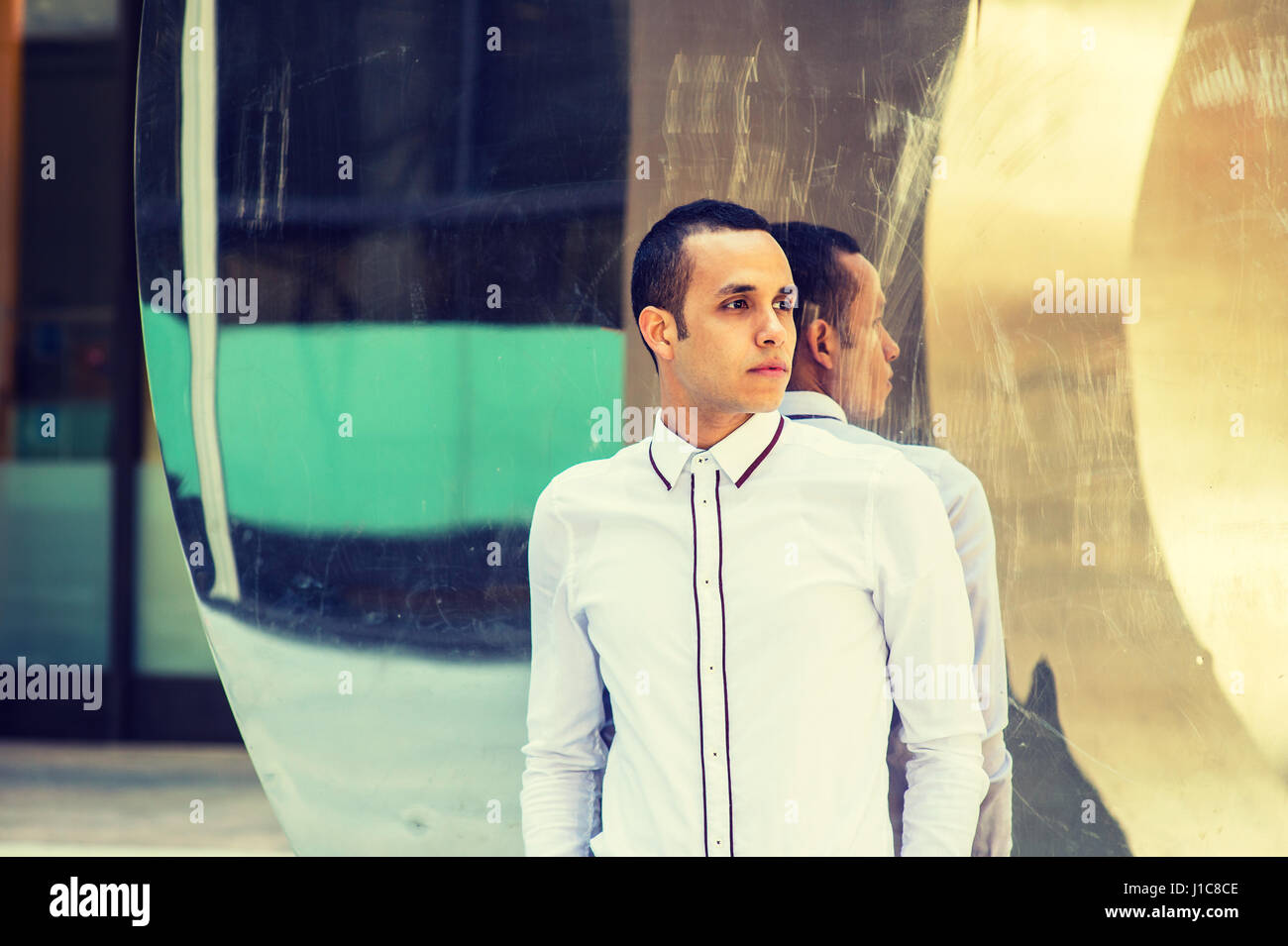 Young American Man wearing white shirt, black pants, standing against metal mirror with reflections in New York, looking away, thinking.  Filtered eff Stock Photo