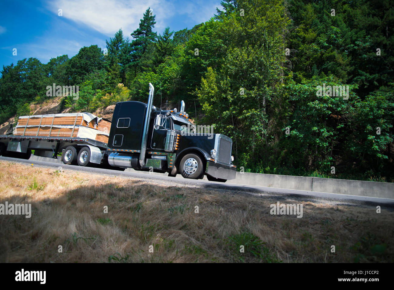 Black stylish semi truck with chrome accessories individual design transports the wood bars on the flat bed trailer moving on the beautiful highway Stock Photo
