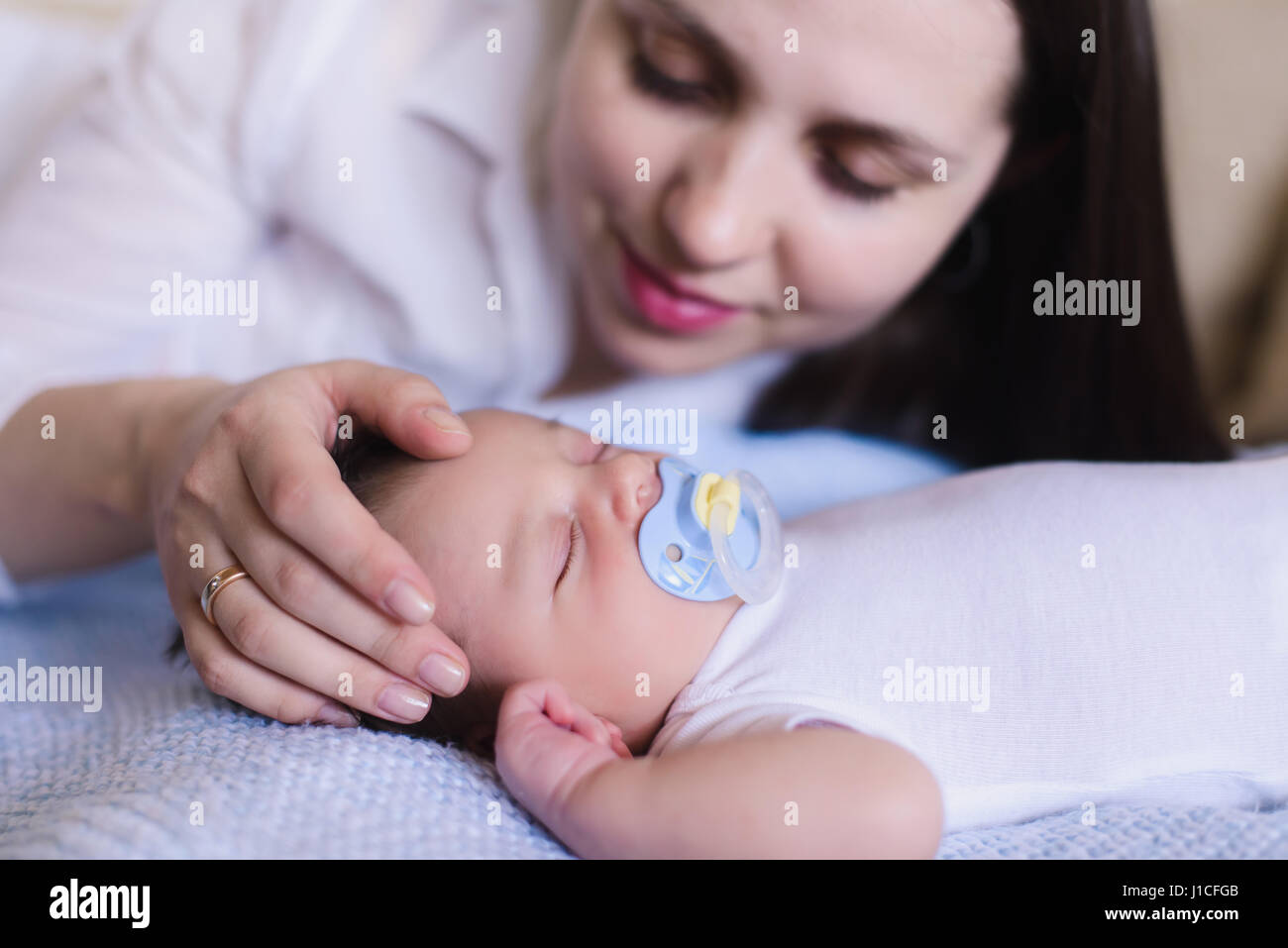 Newborn baby boy with mother. Mom gently stroking the babys head. Baby sweet sleeping on a white bed. Child with a pacifier in his mouth. Stock Photo