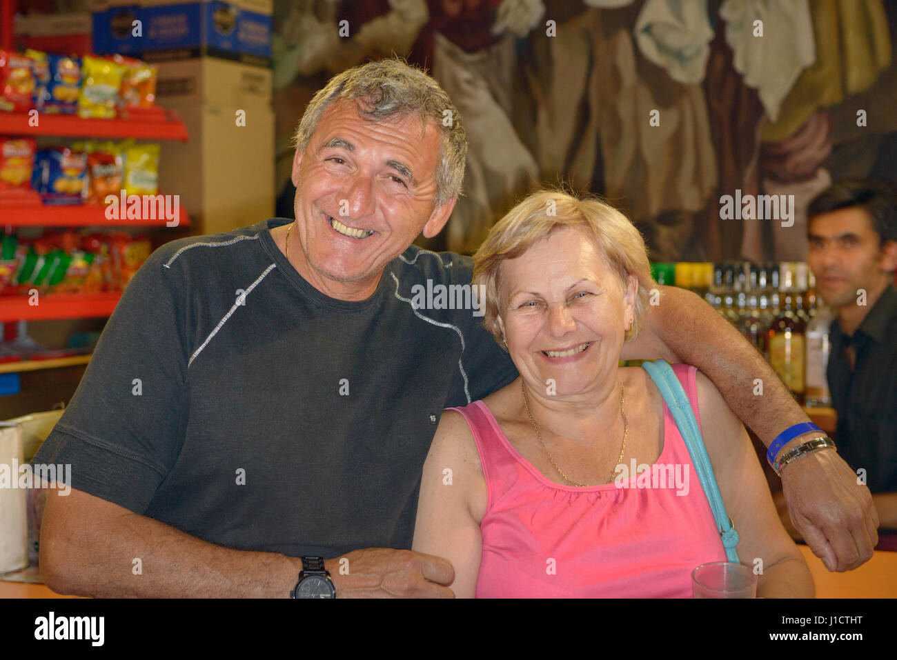 Smiling aged man is embracing the laughing elderly woman shoulders in bar. Stock Photo