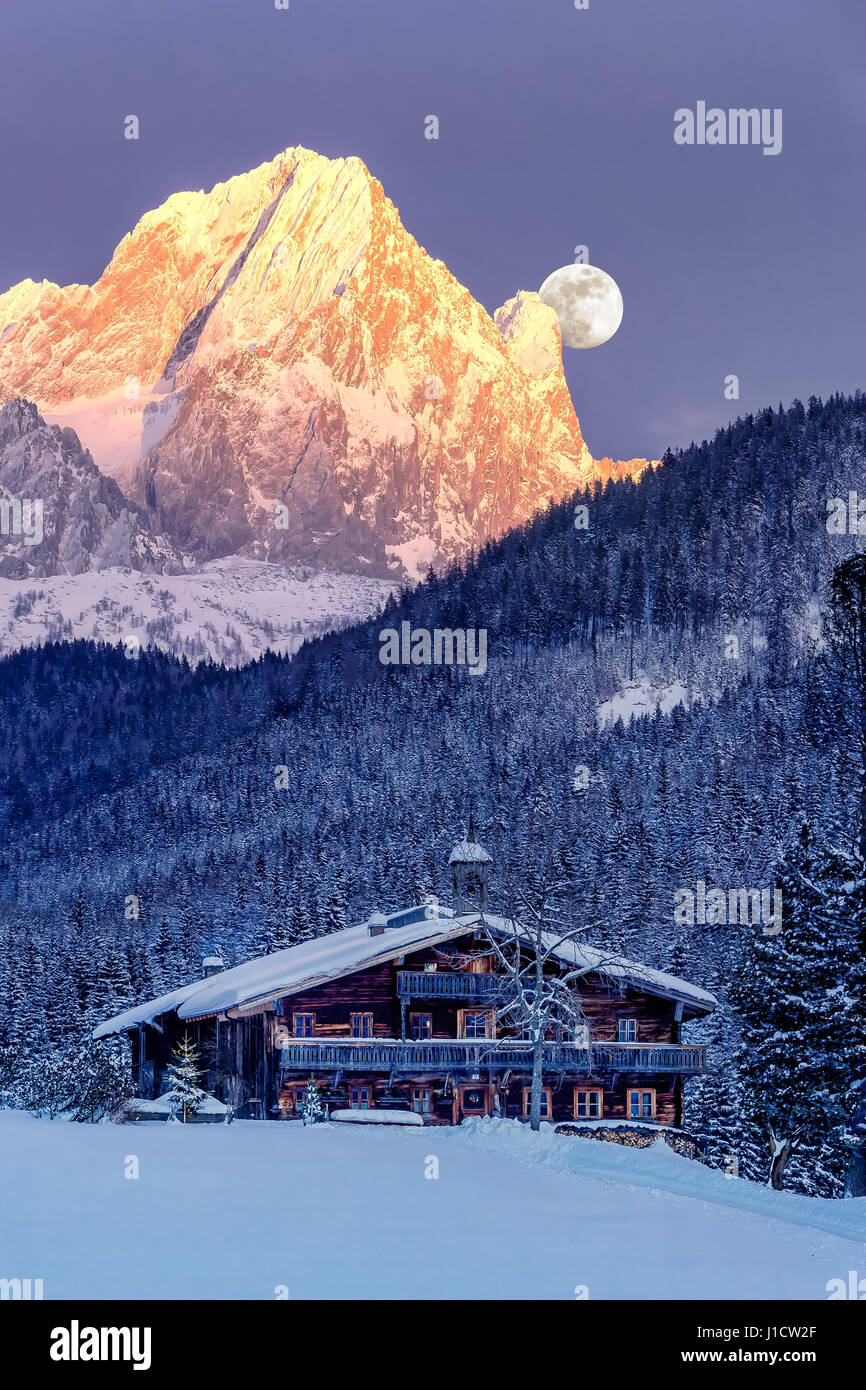 Full moon rises behind the snow-caped Alps with old, traditional farm house in the foreground. Stock Photo