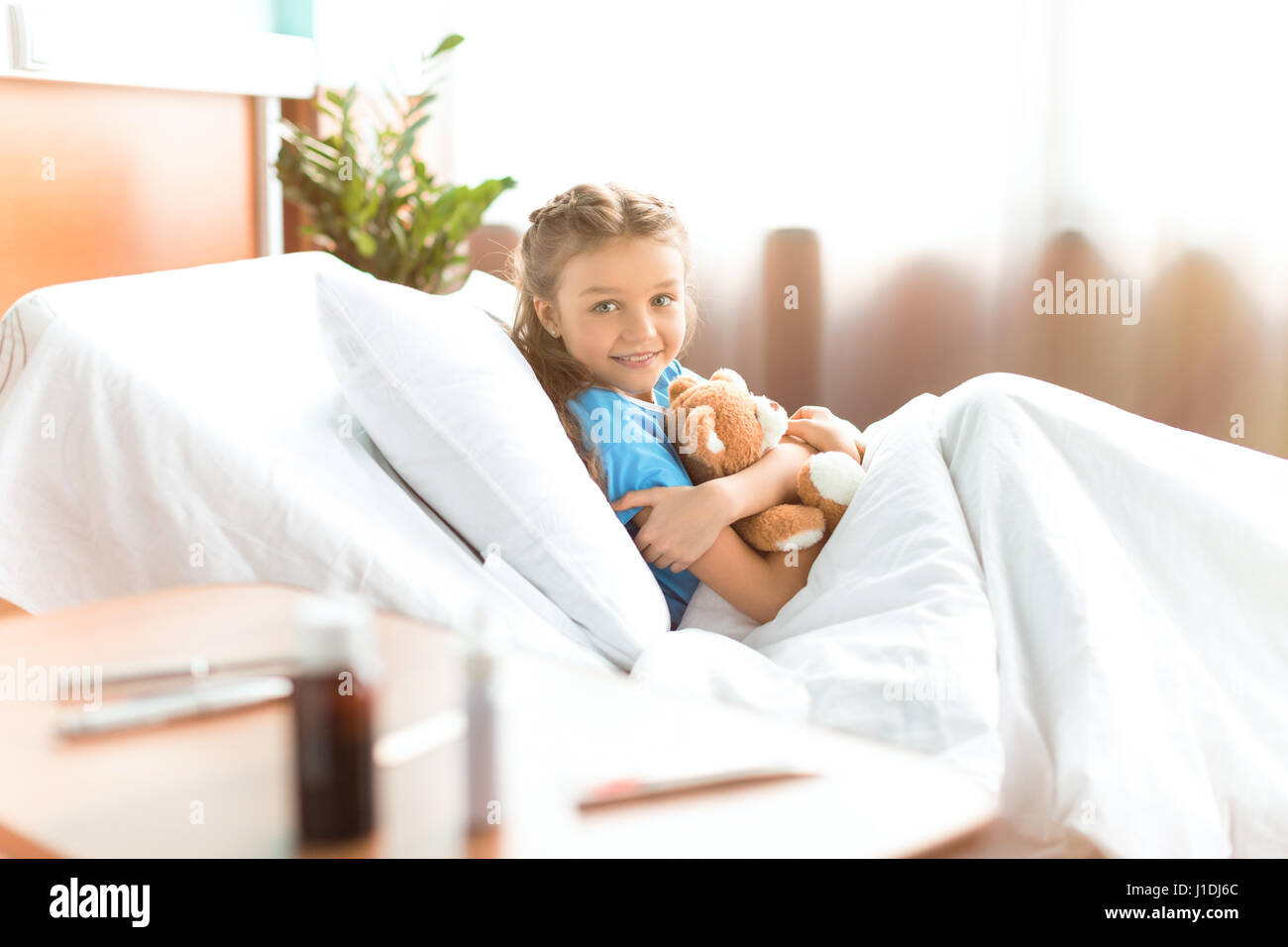 Cute little girl lying in hospital bed with teddy bear and smiling at camera Stock Photo