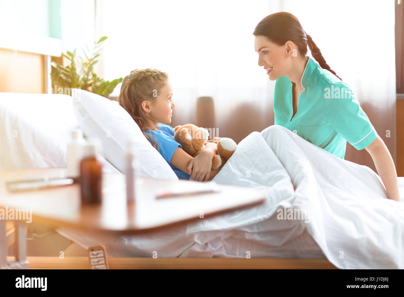 Cute little girl lying in hospital bed with teddy bear and talking with smiling nurse Stock Photo