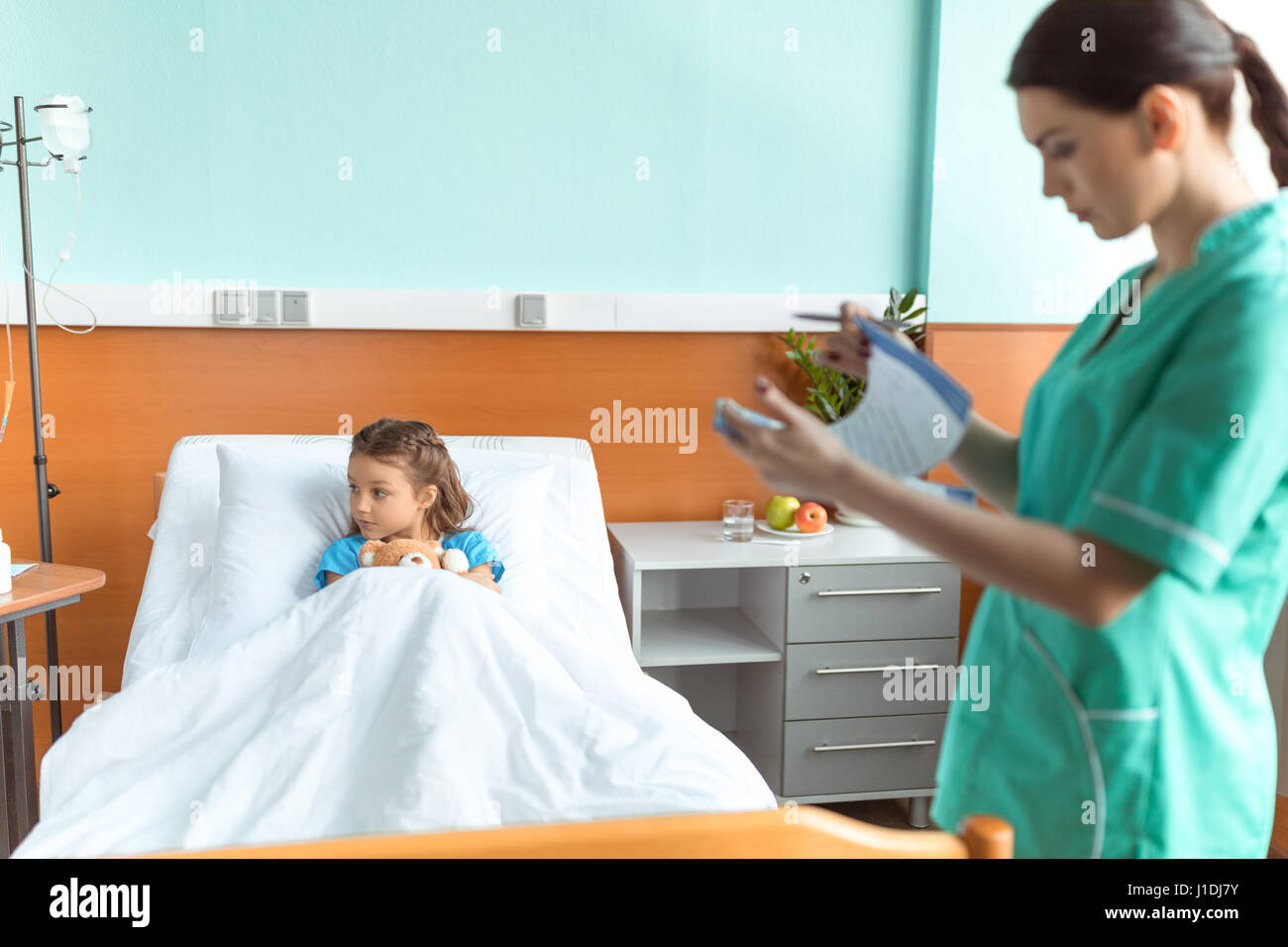 Nurse holding clipboard while standing near sick little girl lying in hospital bed Stock Photo