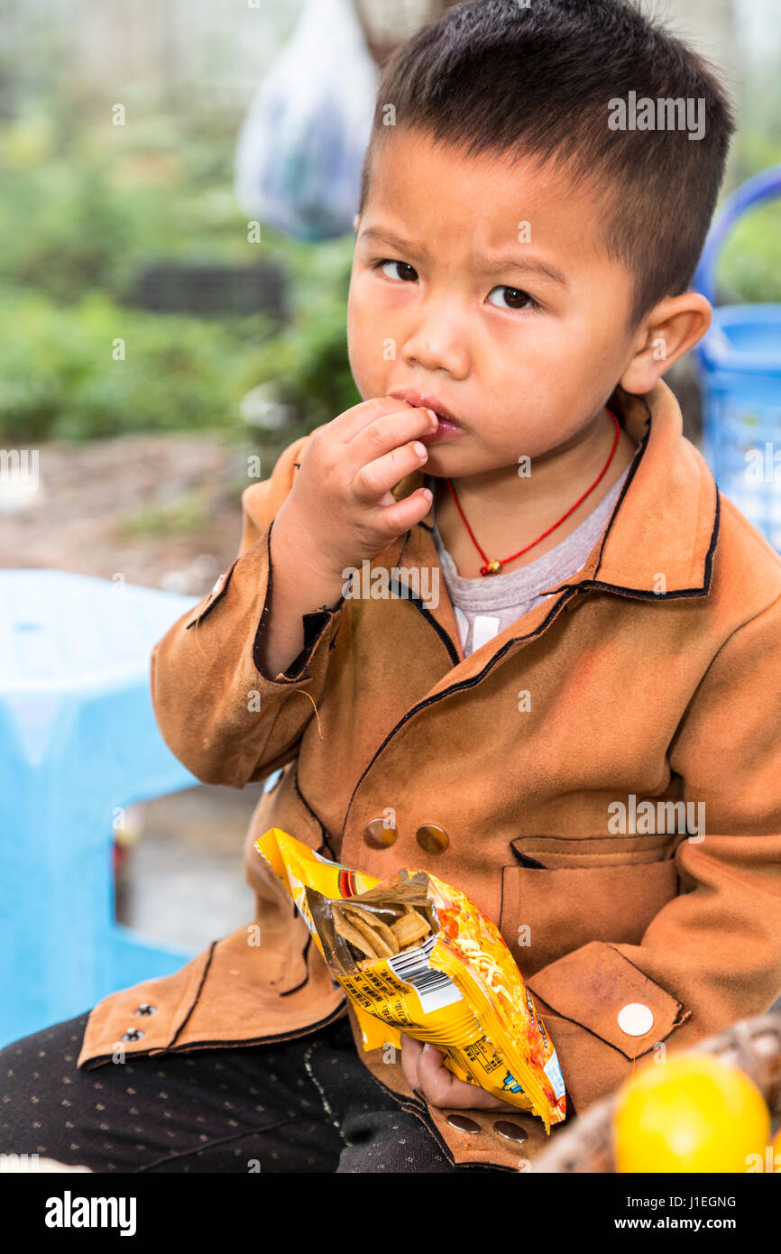 Guizhou, China.  Young Shui Boy Eating a Snack at a Roadside Rest Stop. Stock Photo