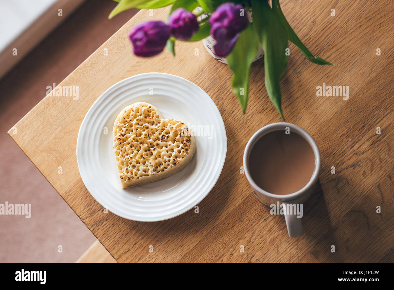 Heart-shaped toasted crumpet on white plate with mug of tea on table with purple tulips. Stock Photo