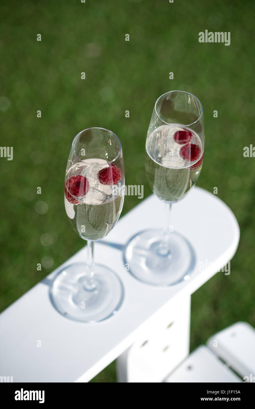 Two glasses of prosecco with raspberries on white garden chair with green grass. Stock Photo