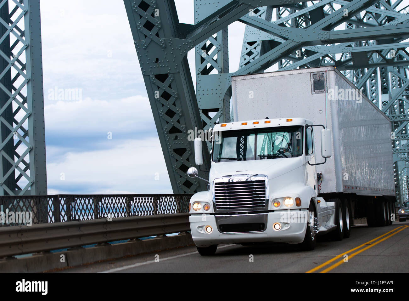 White large commercial semi truck with a trailer carrying commercial industrial loads moving on the highway passing over the bridge with big truss Stock Photo