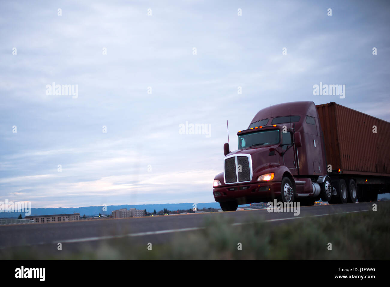 Maroon classic big rig semi truck with headlights transports container on the road running along the industrial and commercial buildings in evening Stock Photo