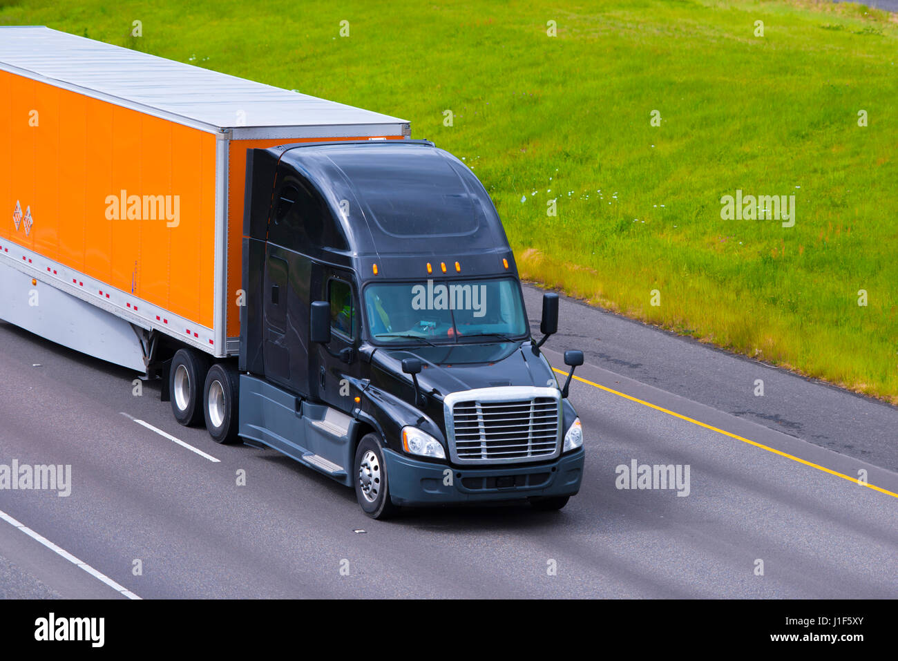 The bright color combination of black modern and showy big rig semi truck and orange dry van trailer with a spoiler on the international road I - 205  Stock Photo