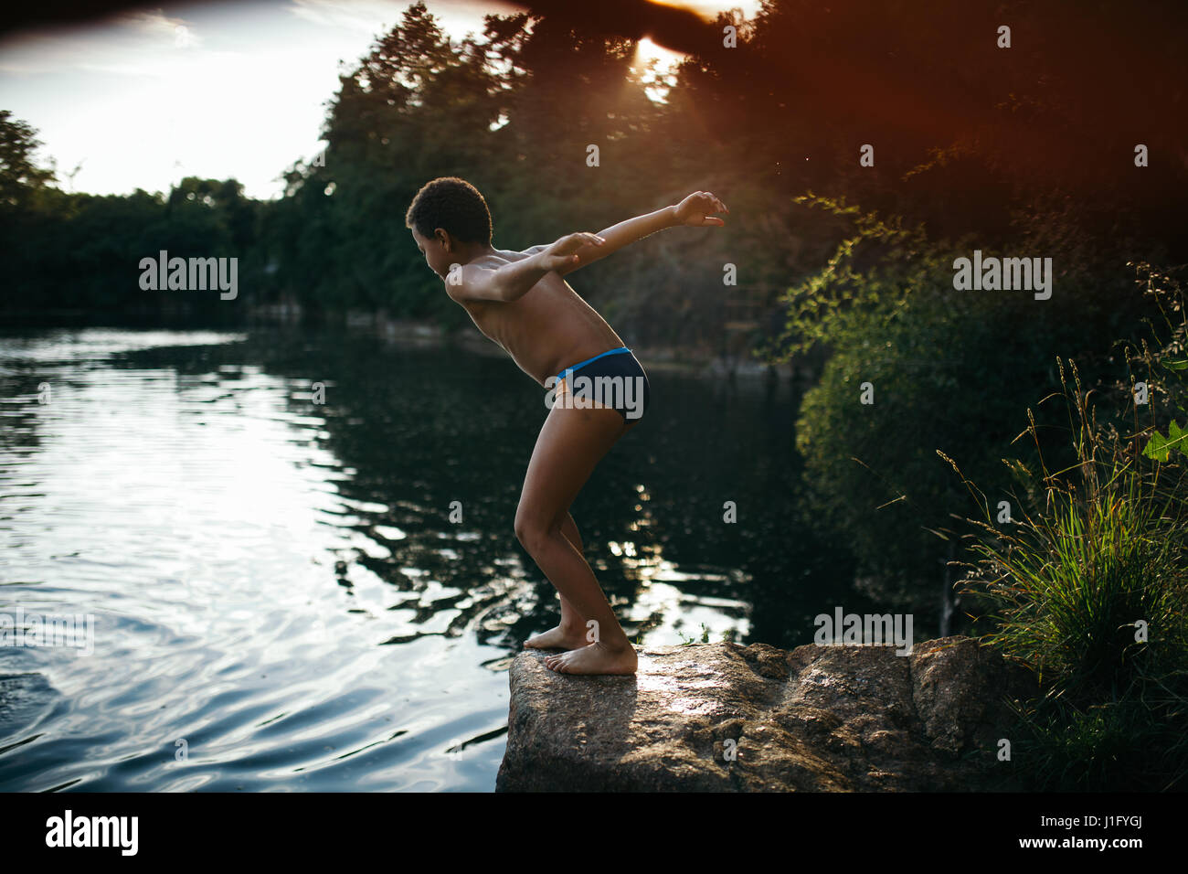 Boy jumping off the cliff into a lake during sunset Stock Photo