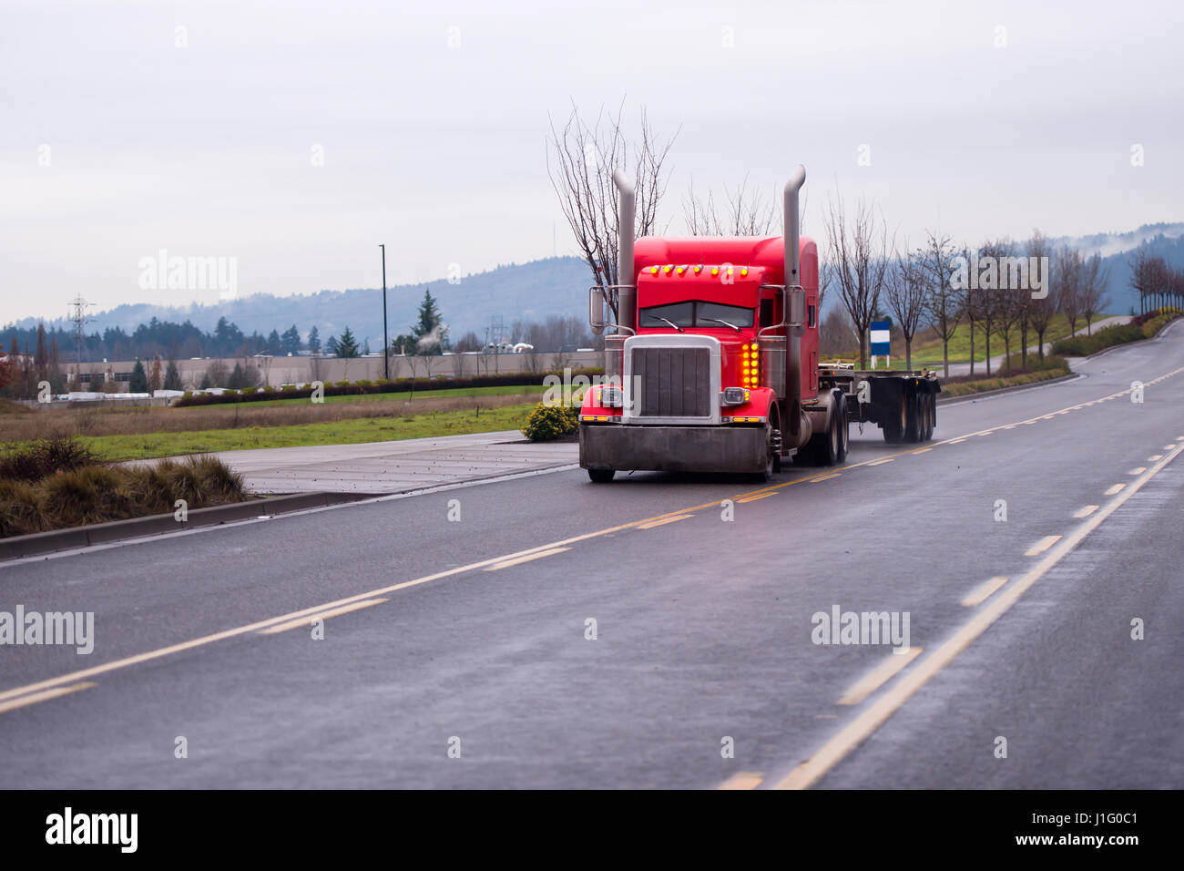 Stylish red classic big rig semi truck with vertical exhaust pipes and the rows of orange sidelights and a trailer on the road with a dividing strips Stock Photo