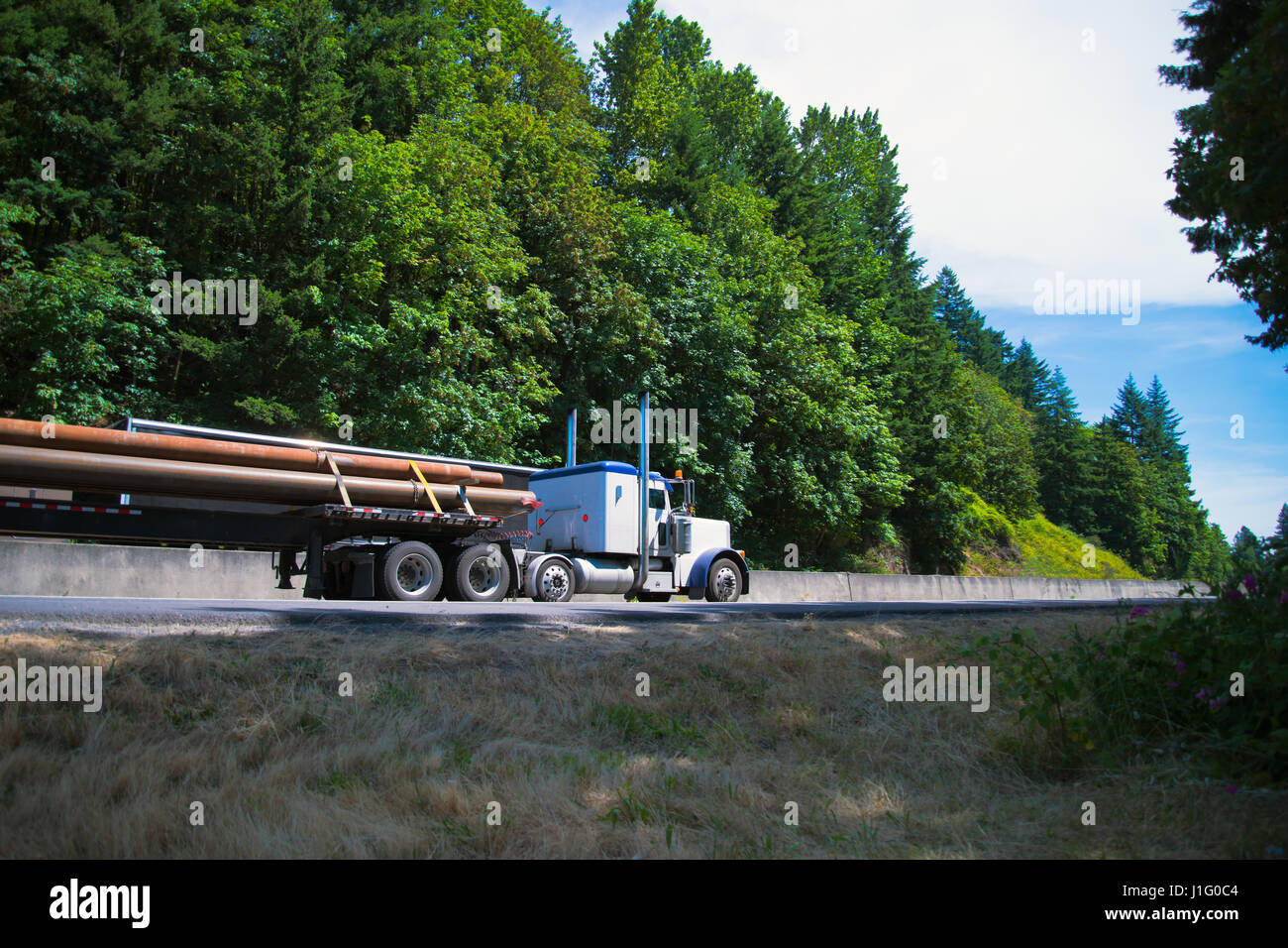 Classic white big rig semi truck with a long oversized load pipe on a flat bed trailer transports cargo on scenic highway surrounded by green trees Stock Photo