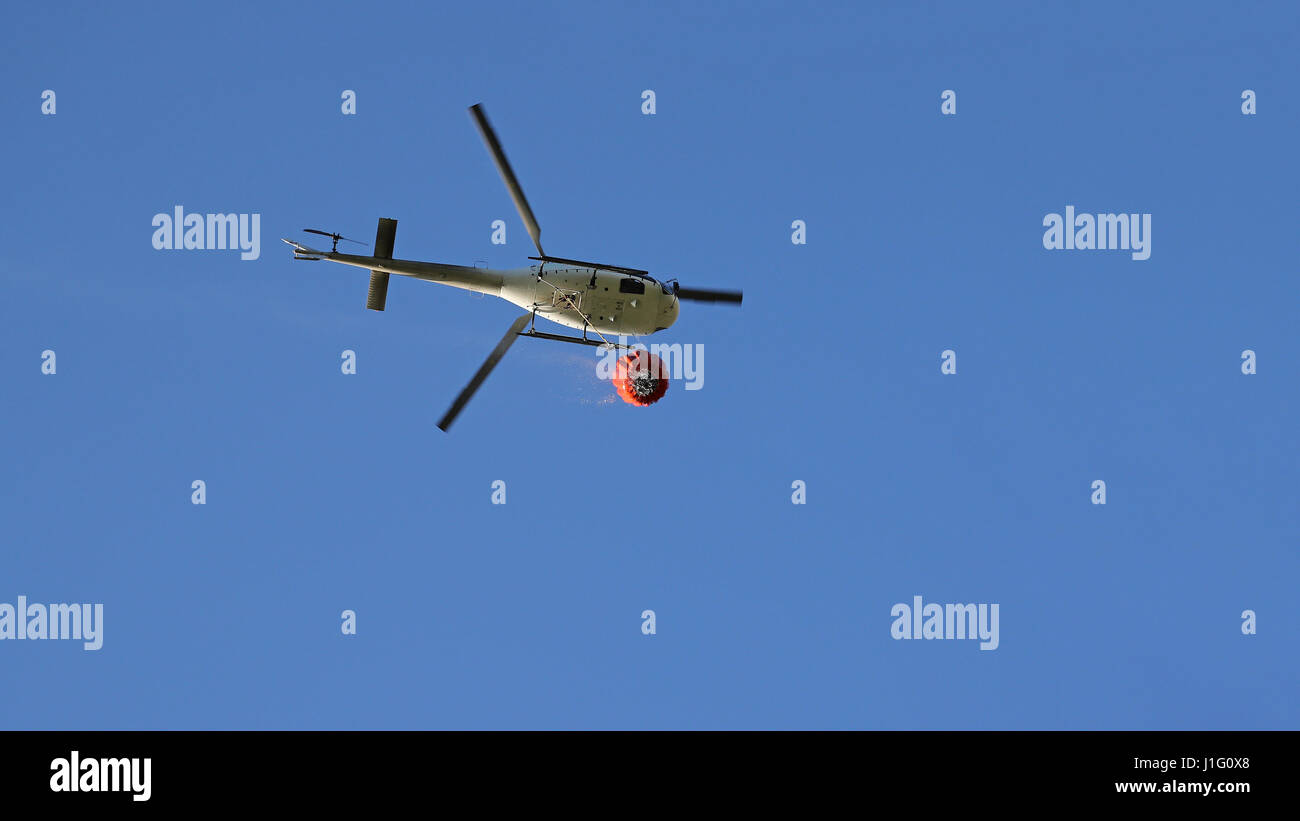 Firefighting helicopter with water bucket and trail of drops seen from below against blue sky