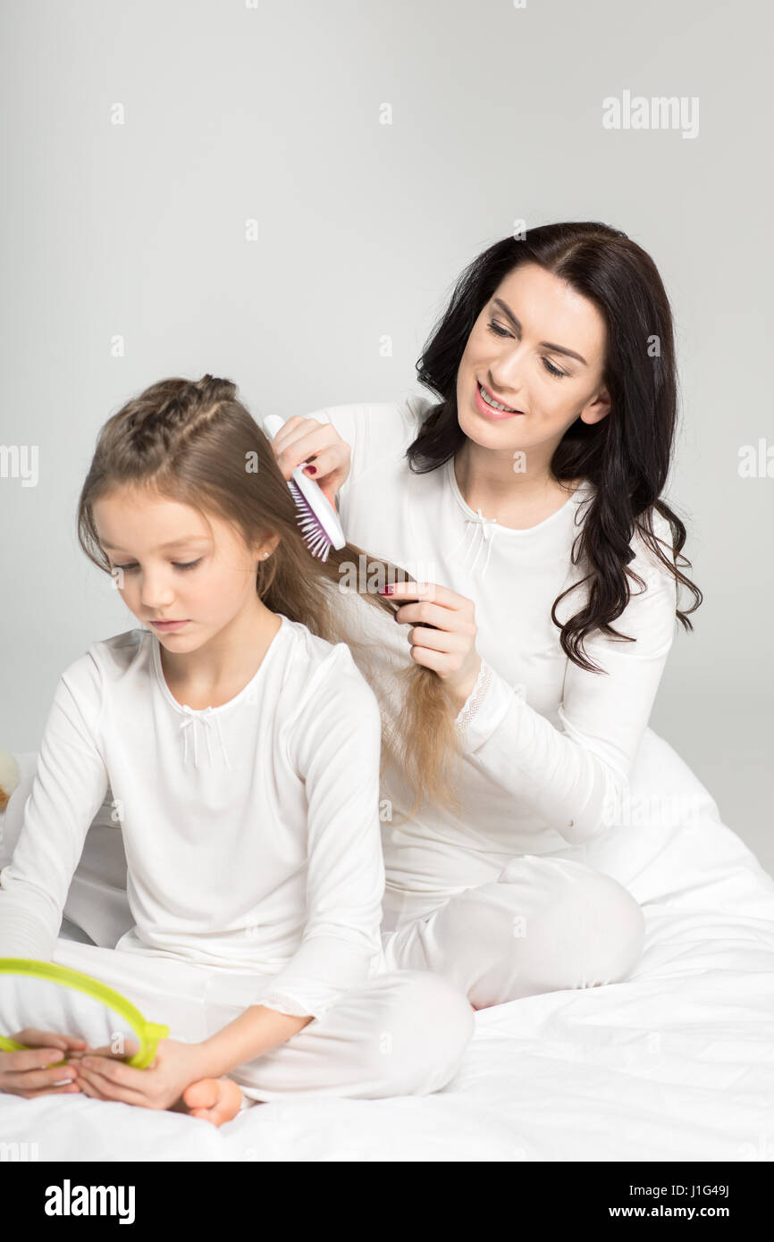 Smiling mother combing hair of cute little daughter with hand mirror Stock Photo