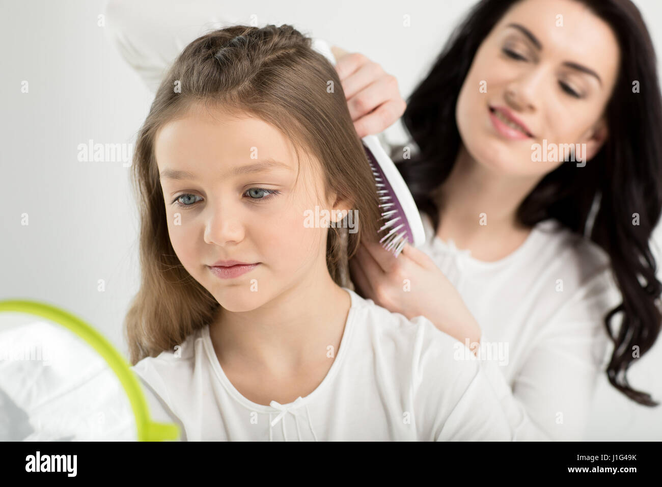 Smiling mother combing hair of cute little daughter looking at hand mirror Stock Photo