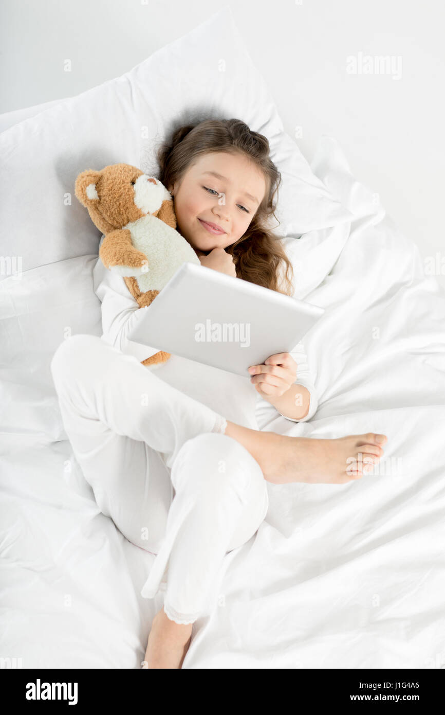 Cute little girl in pajamas using digital tablet while lying in bed ...
