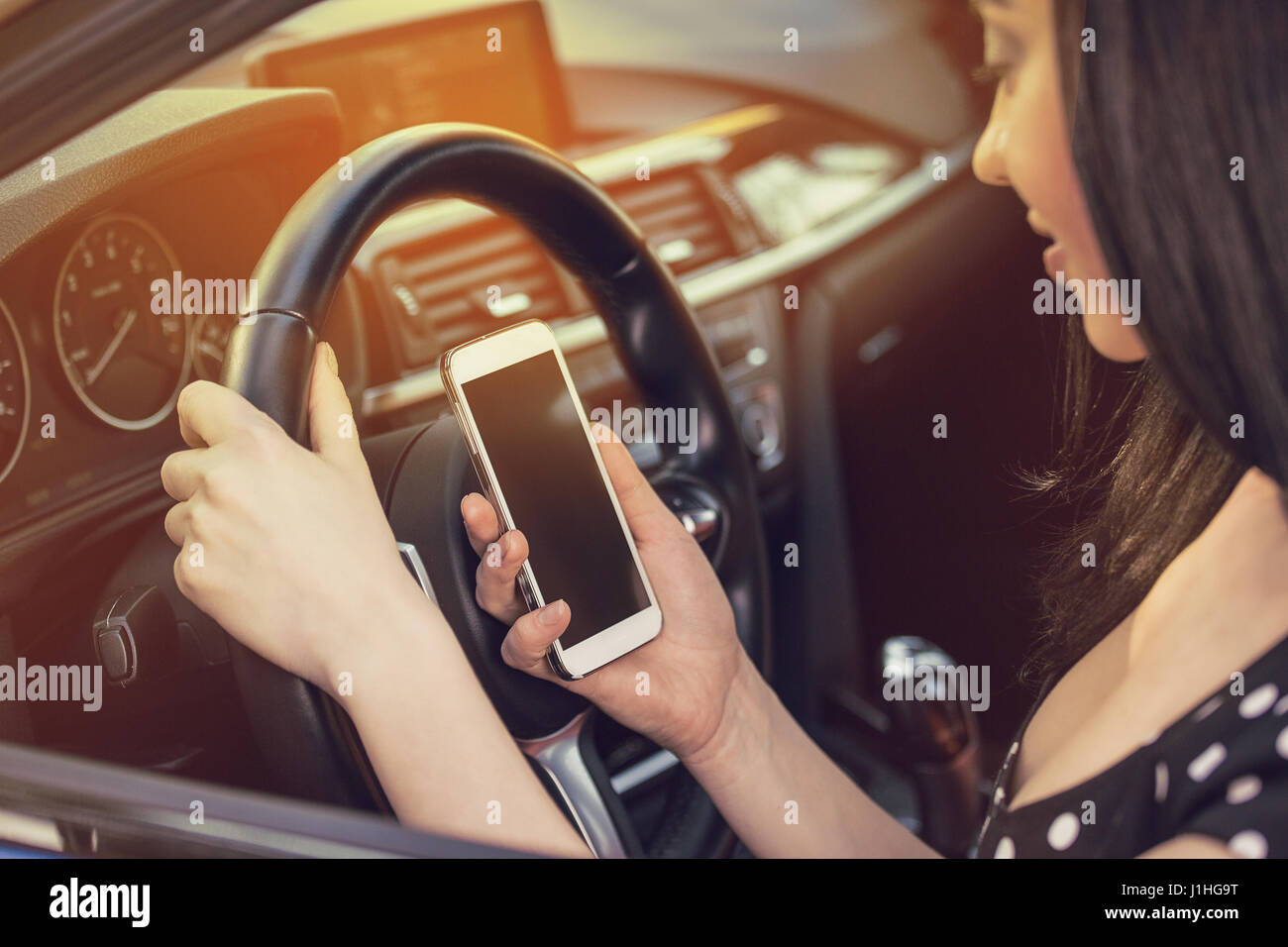 Young woman looking at her smartphone while driving a car on a sunny day with sun light coming through windshield Stock Photo