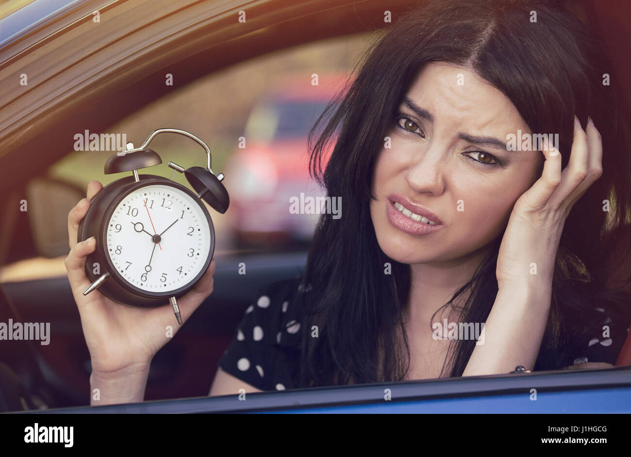 Worried woman sitting inside her car showing alarm clock running late